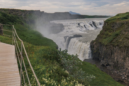 Gullfoss waterfall flows into a rugged gorge with mist rising, surrounded by green vegetation, rocky cliffs, a wooden walkway, and a cloudy sky.の写真素材