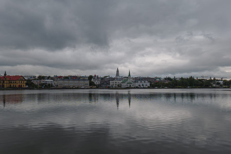 View of Reykjavik, Iceland, featuring Tjornin pond, Hallgrimskirkja spire, Frikirkjan church, and colorful traditional architecture under a cloudy sky.の写真素材