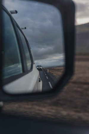A car's side mirror reflects a vehicle following on a narrow road in barren Icelandic terrain. Overcast skies and rugged landscape create a moody scene.の写真素材
