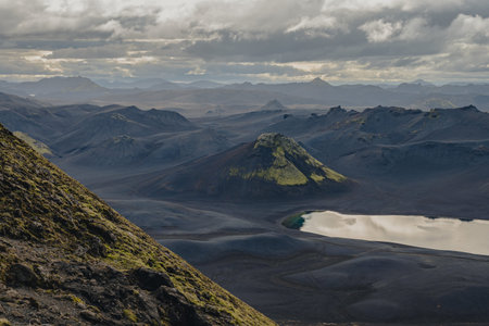A volcanic terrain in Iceland features dark barren land, mossy hills, a reflective lake in the foreground, and distant mountains under cloudy skies.の写真素材