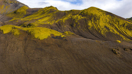 A volcanic terrain with dark slopes, vibrant green moss on ridges, and smooth hills showing erosion patterns under a partly cloudy sky.の写真素材