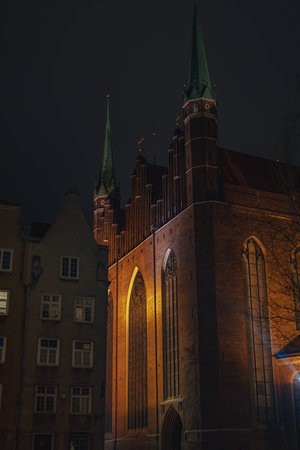 A Gothic style brick church in Gdansk, Poland, with tall spires and arched windows lit by warm lights, surrounded by classic residential buildings at night.の写真素材