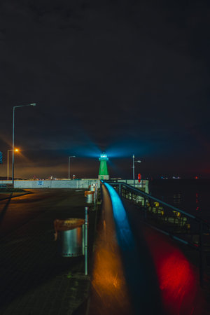 A green lighthouse emits a bright beam at the end of a pier in Gdansk. Reflections of red and blue lights appear on the metallic railing under a cloudy sky.の写真素材