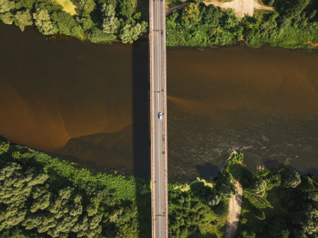 A straight bridge crosses a calm, brownish river surrounded by lush green vegetation. A single vehicle is on the bridge, with trees lining the riverbanks.の写真素材