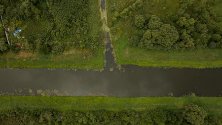 An aerial view of a narrow river bordered by grassy banks and dense vegetation near Berezivka or Minsk, with a small blue roofed structure visible.の写真素材