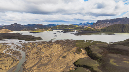 Aerial view of Landmannalaugar in Iceland, featuring a large lake, multicolored rhyolite mountains, braided rivers, and snow capped peaks under a cloudy sky.の写真素材