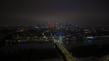 Aerial night scene of central Warsaw shows Swietokrzyski Bridge over the Vistula River, the Palace of Culture and Science in red, Warsaw Spire, and Varso Tower under clouds.の写真素材