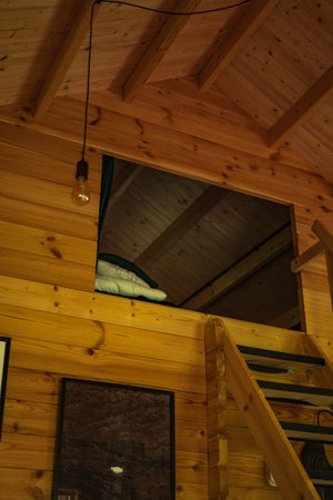 Cozy wooden loft interior in Lithuania shows warm pine planks, exposed rafters, a minimalist hanging bulb, and a narrow stair to an open sleeping loft with folded bedding.の写真素材