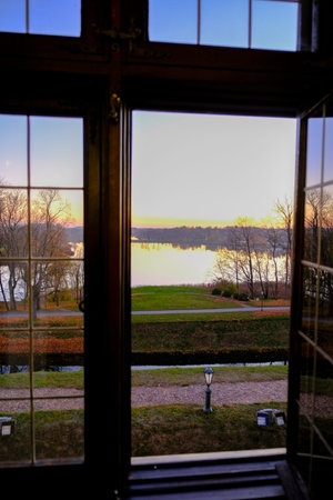 A mullioned window frames a lakeside scene at sunset. A cobblestone path, a vintage lamp post, and a...