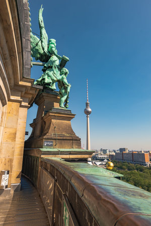 A daytime view from Berlin Cathedral walkway shows a verdigris angel statue, warm light on sandstone, green patina balustrade, and the Fernsehturm over central Berlin.の写真素材