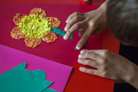 The young man at the table engaged in creativity. Children's hands make the application. The boy's hands close up make a flower out of paper. Children's entertainment. The creativity of paper.の写真素材