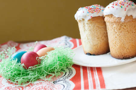 Easter cakes on a plate, next to the bright colored Easter eggs. Painted chicken eggs are in...