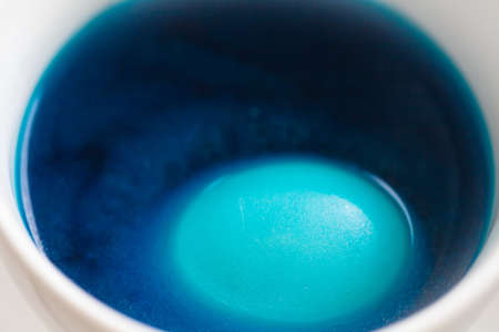 The process of coloring Easter eggs. Eggs are immersed in liquid with paint. White mugs with colored liquid stand on the windowsill. Blue spot.の写真素材