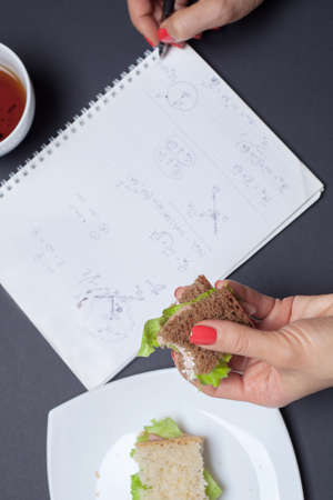 Girl's hand holding a sandwich and write formulas in Notepad. Mug of tea next to a plate of sandwiches. Girl at work eating bruschetta. Food intake during the study.の写真素材