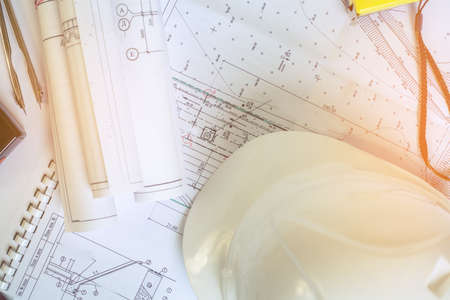 The sun illuminates the table with documents. A metal compass lies on the drawings. Accessories construction engineer. White safety helmet lies on the drawings. Coils projects on the documents.の写真素材