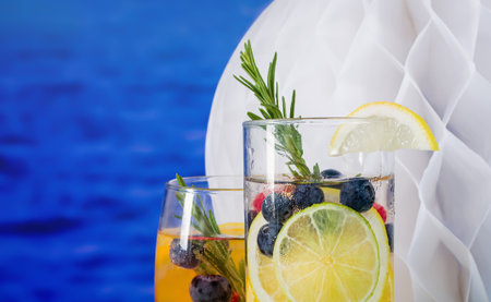A portion of a drink with a slice of lemon and a sprig of rosemary against the backdrop of the sea