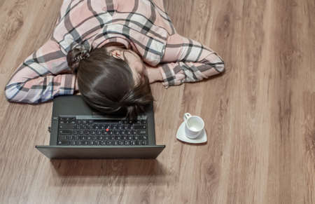 View from above. Girl sleeps with her head resting on a computer keyboard. Wooden bright yellow floor. A white empty coffee cup is nearby. Concept of the problems associated with working from home.の写真素材