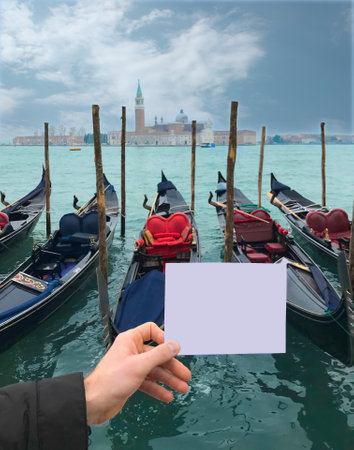 A man's hand holds a small blank sheet of paper against the background of the mooring of Venetian gondolas. Blank for an inscription, template, mock-upの写真素材