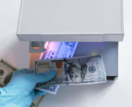 A hand in a blue glove holds three banknotes for verification. The ultraviolet lamp of the currency...