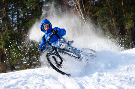Cyclist extreme riding mountain bike in flying snow near winter forest in sunny cold dayの写真素材