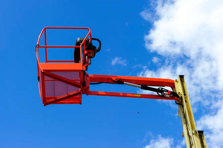 Hydraulic lift platform with bucket of construction vehicle painted in orange and beige colors with white clouds and blue sky on backgroundの写真素材