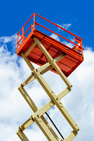 Scissor lift platform with hydraulic system at maximum height range painted in orange and beige colors, large construction machine, heavy industry, white clouds and blue sky on backgroundの写真素材