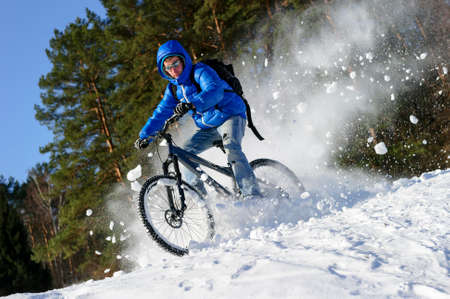 Cyclist riding bike, extreme winter cycling near snowbanks of snowy mountain slope, cross country biking near forest in cool sunny dayの写真素材