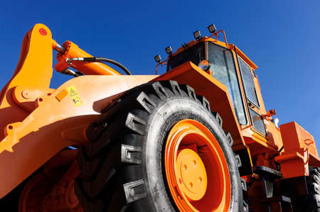 Bulldozer, huge orange powerful construction machine with hydraulic piston of scoop and black wheels, heavy industry, bottom view, blue sky on backgroundの写真素材