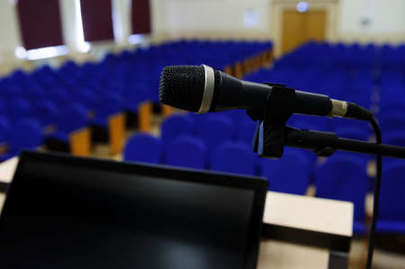 Microphone in conference hall, auditorium for business meeting and presentation with rostrum and display for speaker and rows of seats for participants and visitors on blurred backgroundの写真素材