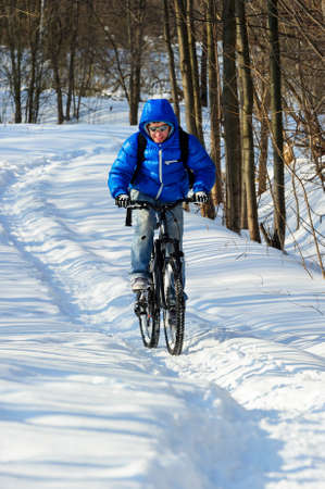 Extreme winter cyclist riding mountain bicycle with effort on rural track among snowbanks near snowy beautiful forest in sunny cold dayの写真素材