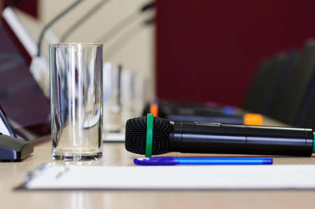 Conference room table for business meeting with microphone and glass for water, pen, paper, monitors, chairs, other microphones and glasses on blurred background, selective focusの写真素材