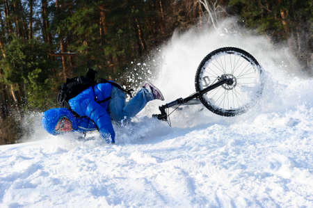 Extreme cyclist falling down from bicycle near snowbanks of mountain slope, winter sport, cross country biking near forest in cool sunny dayの写真素材