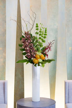 Floral arangement with Cymbidium and James Storey orchids, Hydrangea, Curly willow, Yellow pin cushion protea and greenery in white cylinder vase, and two beige chairsの写真素材