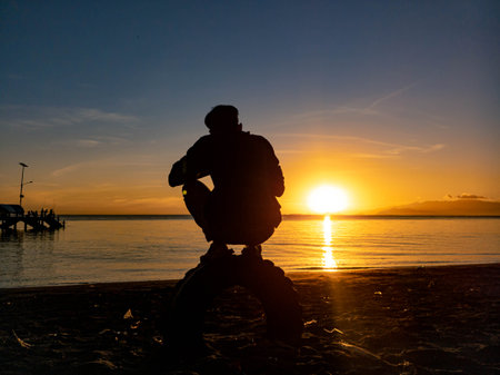 Silhouette of a man sitting on the beach at sunset.の写真素材