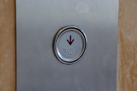 Close up view of an elevator button with downward arrow and braille dots on metal panel.