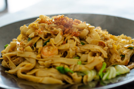 Close-up shot of hot Indonesian Kwetiau Goreng, stir-fried flat rice noodles with shrimp, egg, vegetables, and crispy shallots served on a black plate.の写真素材