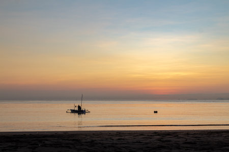 Fisherman silhouette on traditional outrigger boat at sunrise over calm ocean with colorful gradient sky.の写真素材