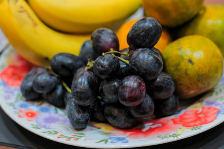 Close-up macro shot of ripe black grapes centered on a patterned floral dish, featuring soft focus on citrus fruits and bananas in the vibrant background.の写真素材