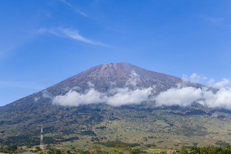 Mount Agung dominating the landscape, partially shrouded in clouds under a clear blue sky with...