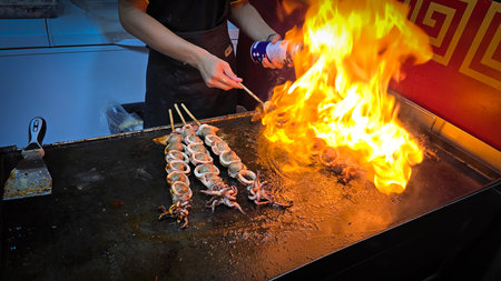cook grills skewered squid over an open flame using a blowtorch, capturing the lively atmosphere of a bustling street food Thai market with bright flames and freshly prepared seafood.の写真素材