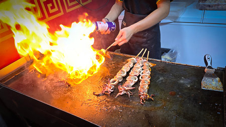 cook grills skewered squid over an open flame using a blowtorch, capturing the lively atmosphere of...