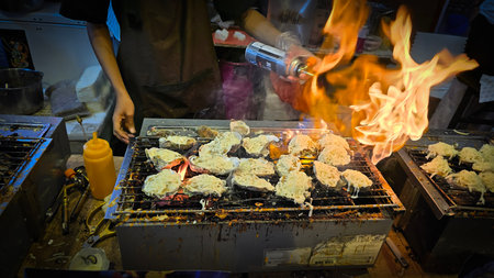 street vendor uses a torch flame to prepare delicious grilled oyster with flair and skill, showcasing the vibrant atmosphere of bustling night Thai markets. Good for recipe bookの写真素材