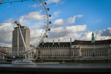 view of the London Eye on the River Thames under a clear blue sky, with clouds and a city skyline along the riverbank, suggesting travel, tourism and urban imagery. Iconic London Eye On The Thames With Bright Sky And Cityscape Panoramaの写真素材
