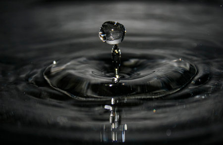 macro capture of a water droplet rising from a narrow column, forming concentric ripples on a dark,...