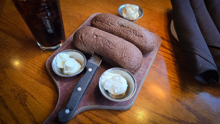 Brown Australian rye loaves rest on a rustic wooden board with small butter cups, a sharp knife, and...