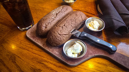 Brown Australian rye loaves rest on a rustic wooden board with small butter cups, a sharp knife, and...