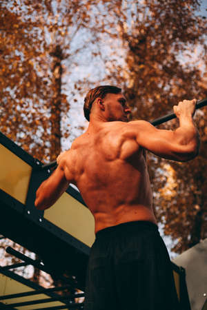 Young sexy athlete man with naked torso doing pull-ups in the park.