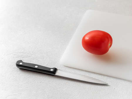 Red ripe tomato on the chopping board and a kitchen knife on the white table. Minimalism. Food conceptの写真素材