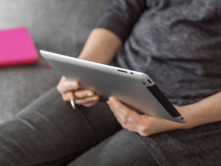 Young girl watching lessons on a tablet sitting on gray sofa. Business woman on quarantine. Home office, online education, remote work, distance learning, stay home conceptの写真素材