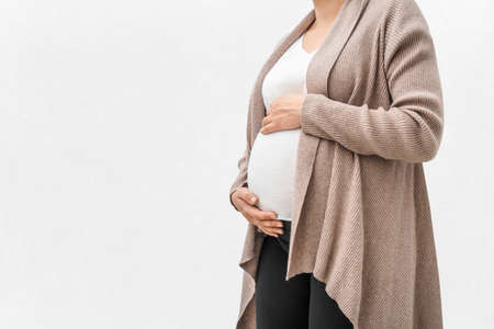 Pregnant woman in woolen beige cardigan holds hands on belly isolated on white background. Pregnancy...